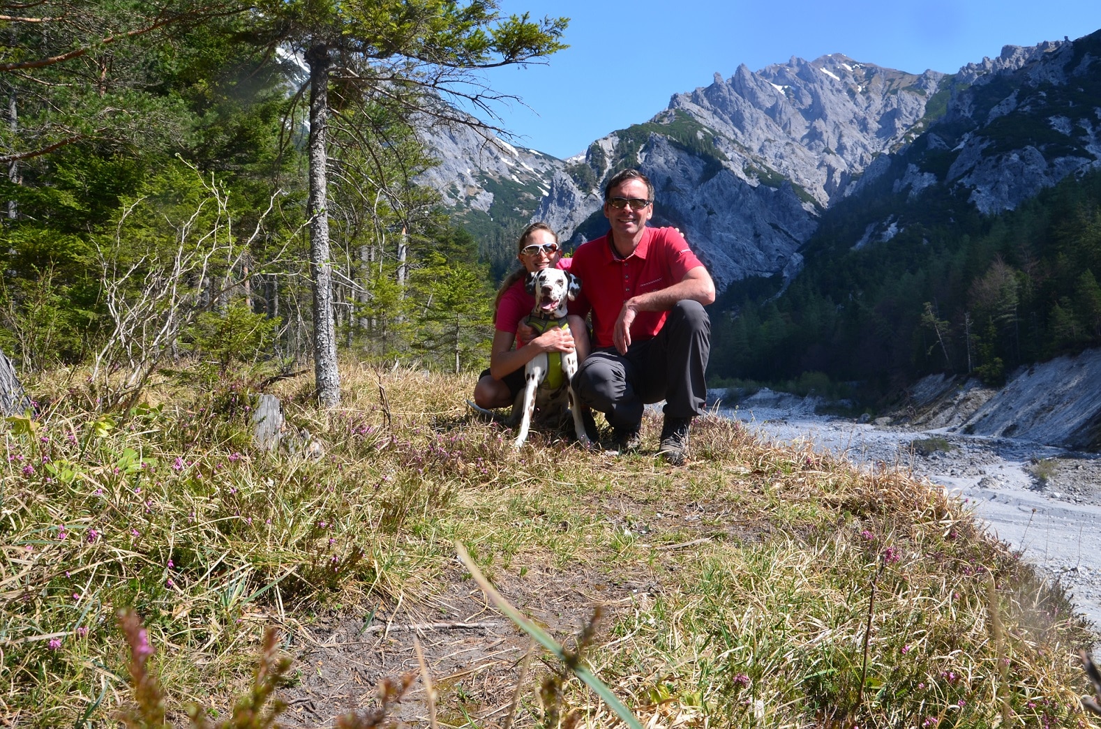 Mann, Frau und Hund sitzen am Waldrand an einem ausgetrockneten Bachbett; im Hintergrund hohe Berge und Felsen