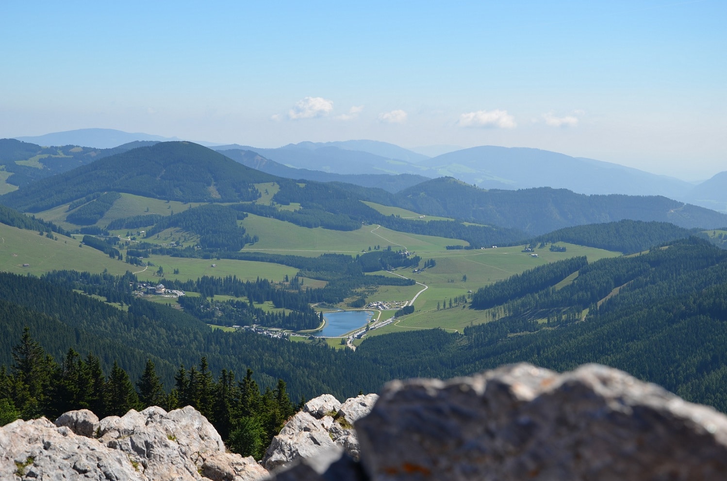 Blick auf sanfte Almen, grüne Hügel, bewaldete Berge und See