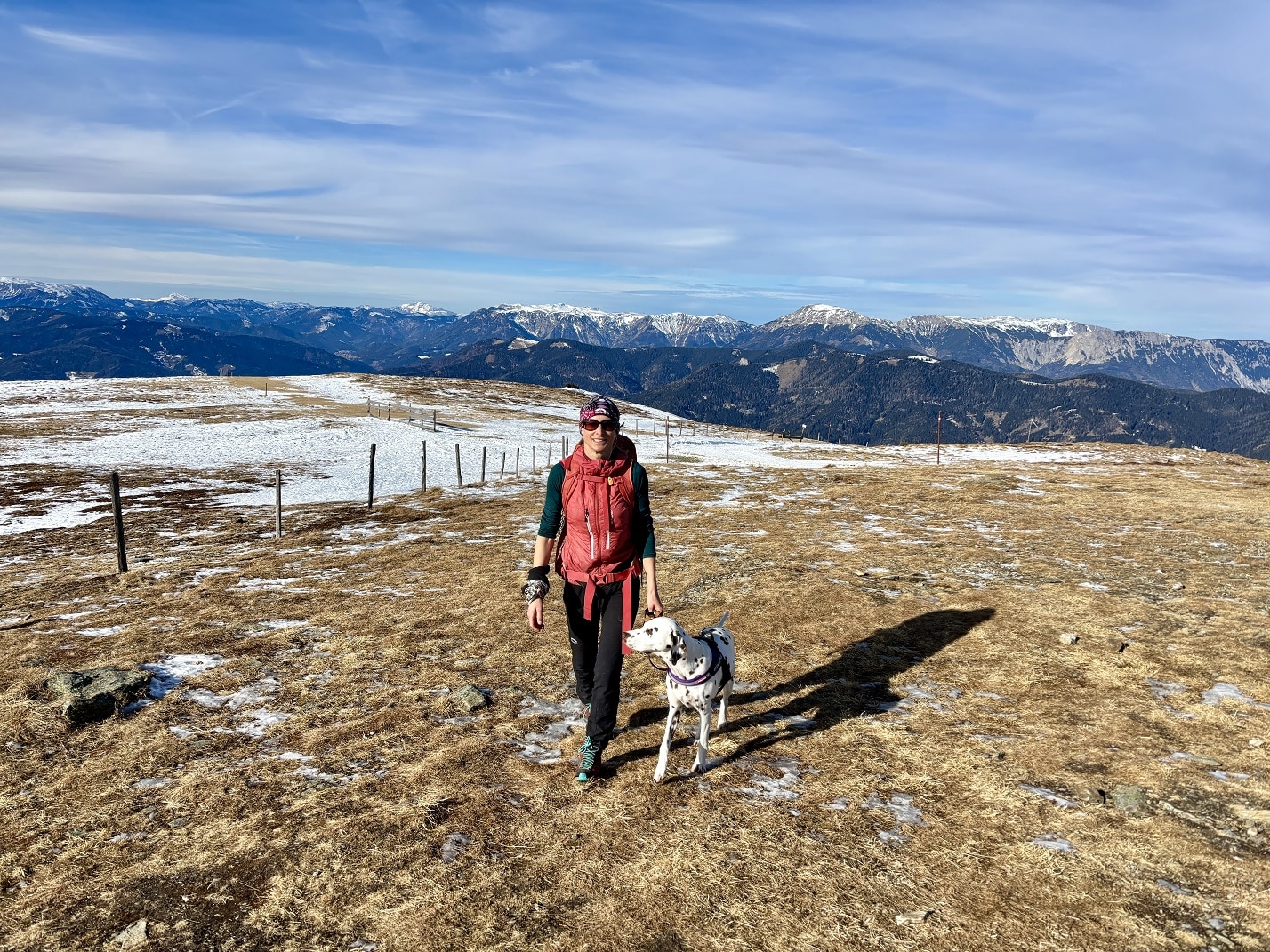 Frau wandert mit Hund auf Almrücken, Berge wenig schneebedeckt