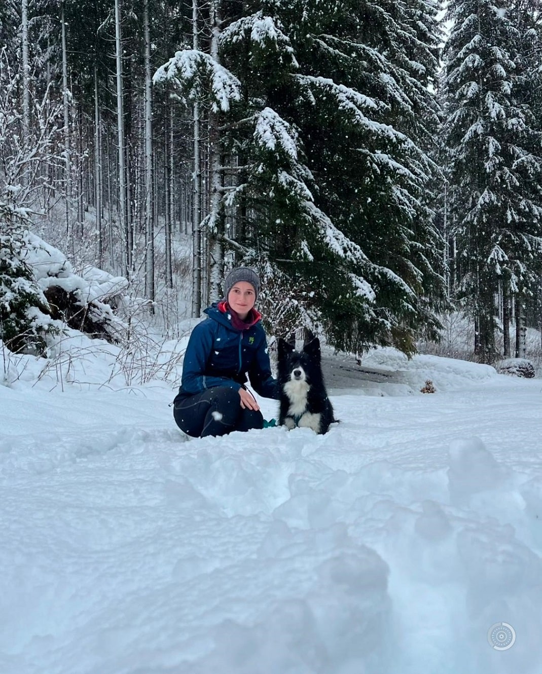 Frau und schwarzer Hund sitzen in verschneiter Landschaft.