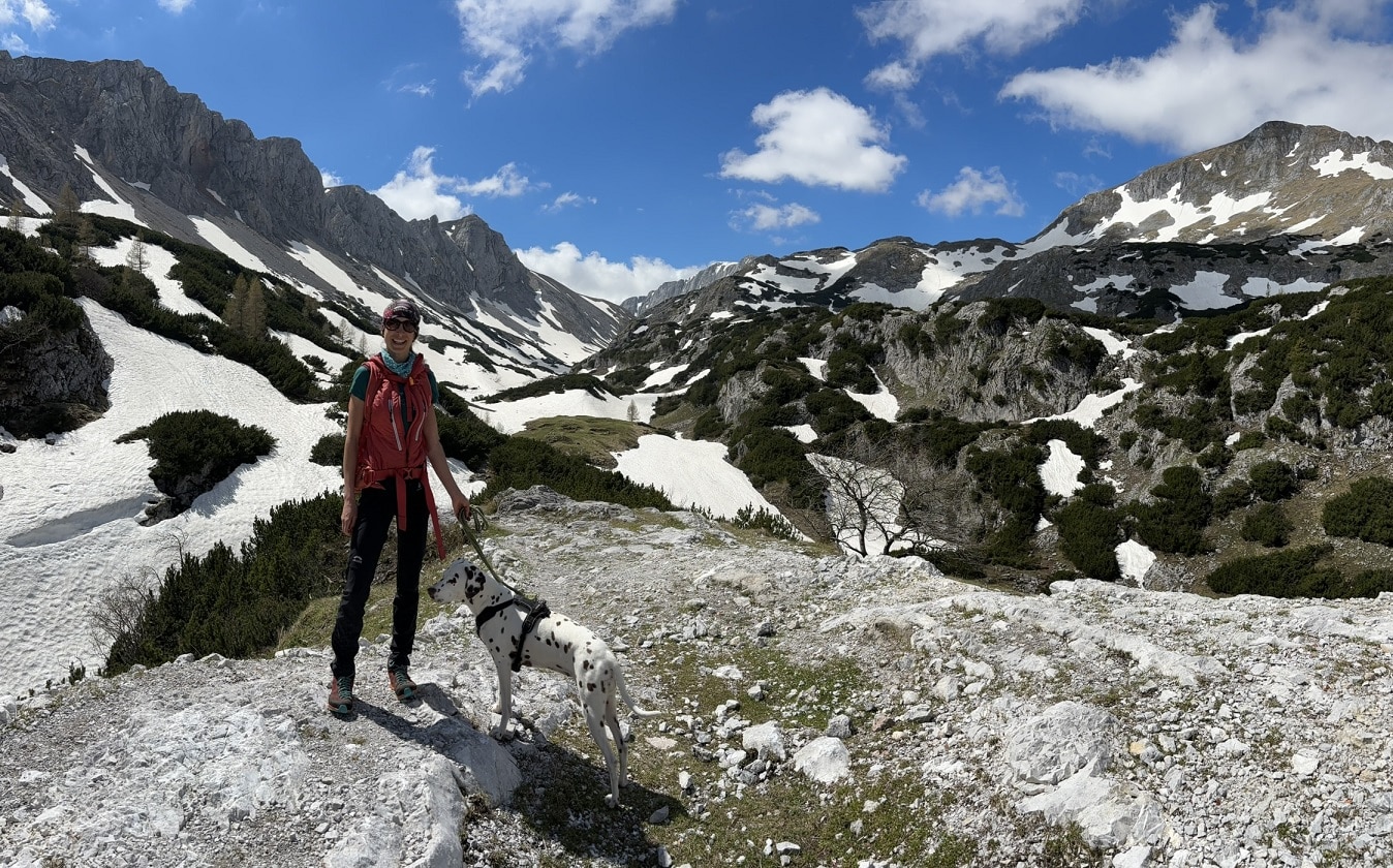 Frau steht mit Hund in alpinem Gelände; Schneeflecken und Kalksteinmassiv im Hintergrund