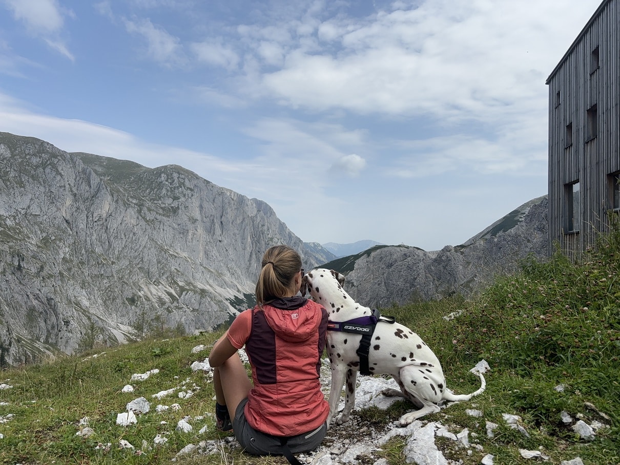 Frau sitzt mit Hund (Dalmatiner) in der Wiese auf einem Berg; am Bildrand ist eine Hütte zu sehen