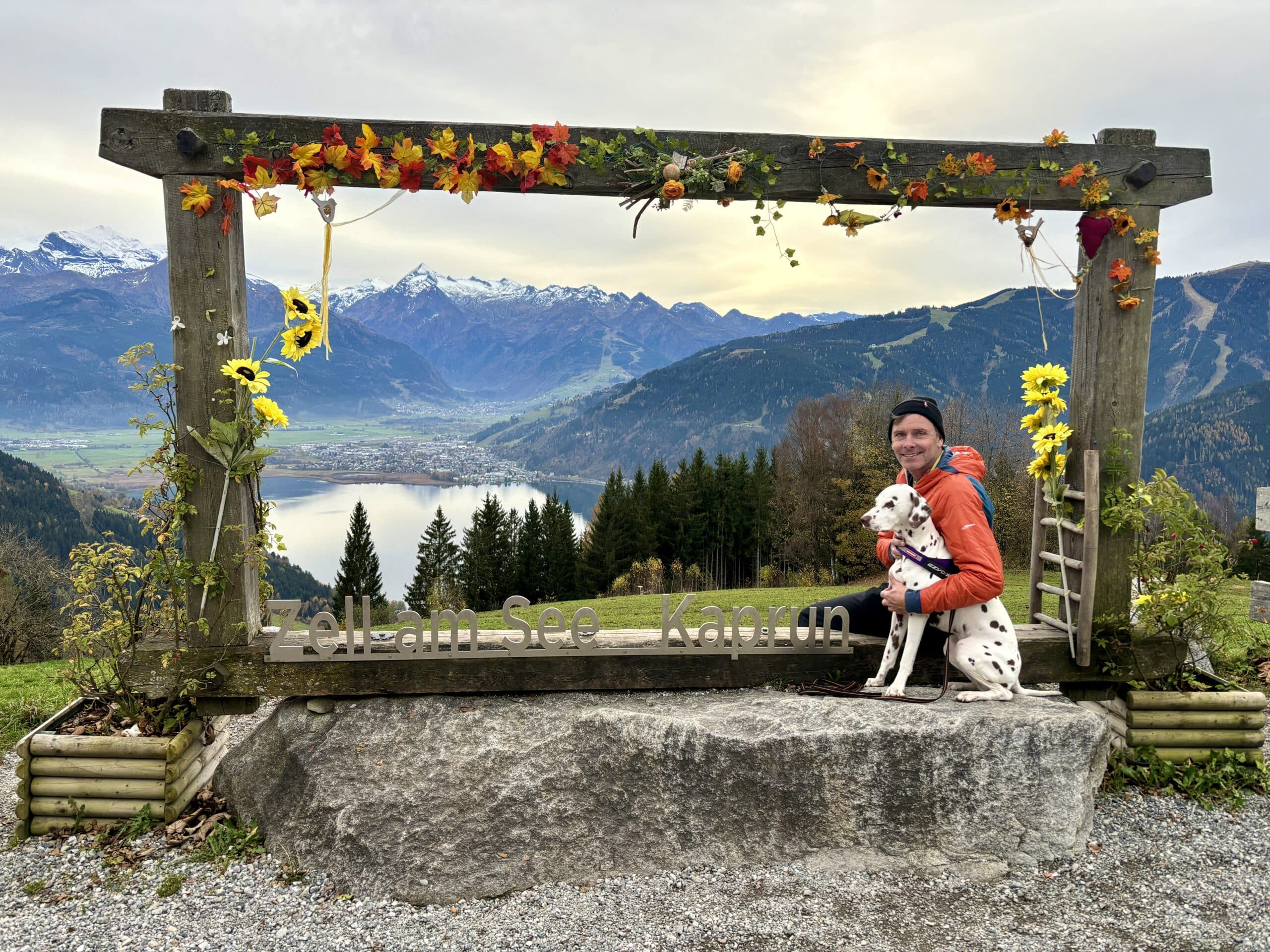 Mann sitzt mit Hund am Berg in einem risengroßen Fotorahmen; im Hintergrund ein See und Berge