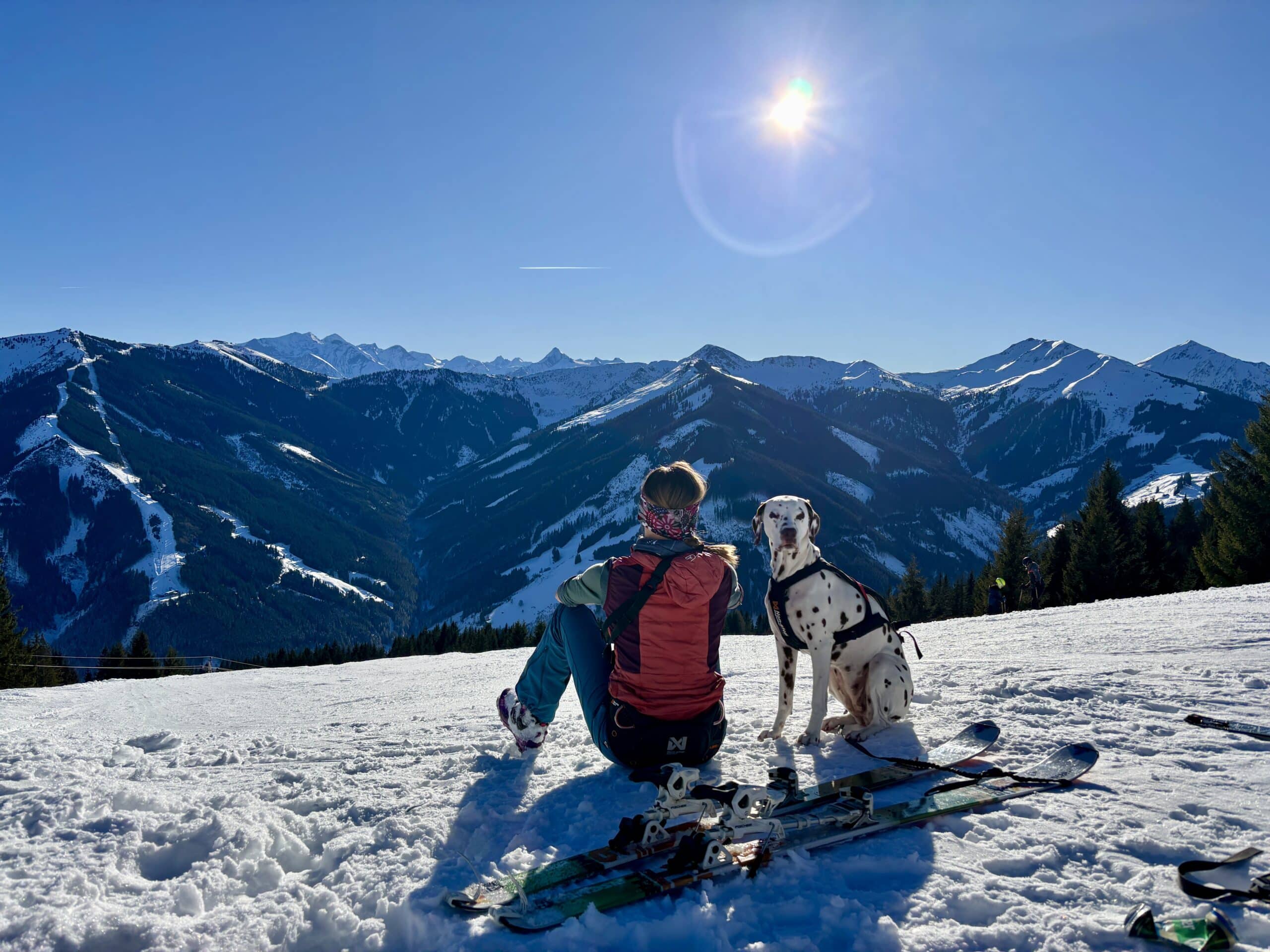 Frau sitzt mit Hund (Dalmatiner) im Schnee und blickt auf verschneite Berge bei Sonnenschein