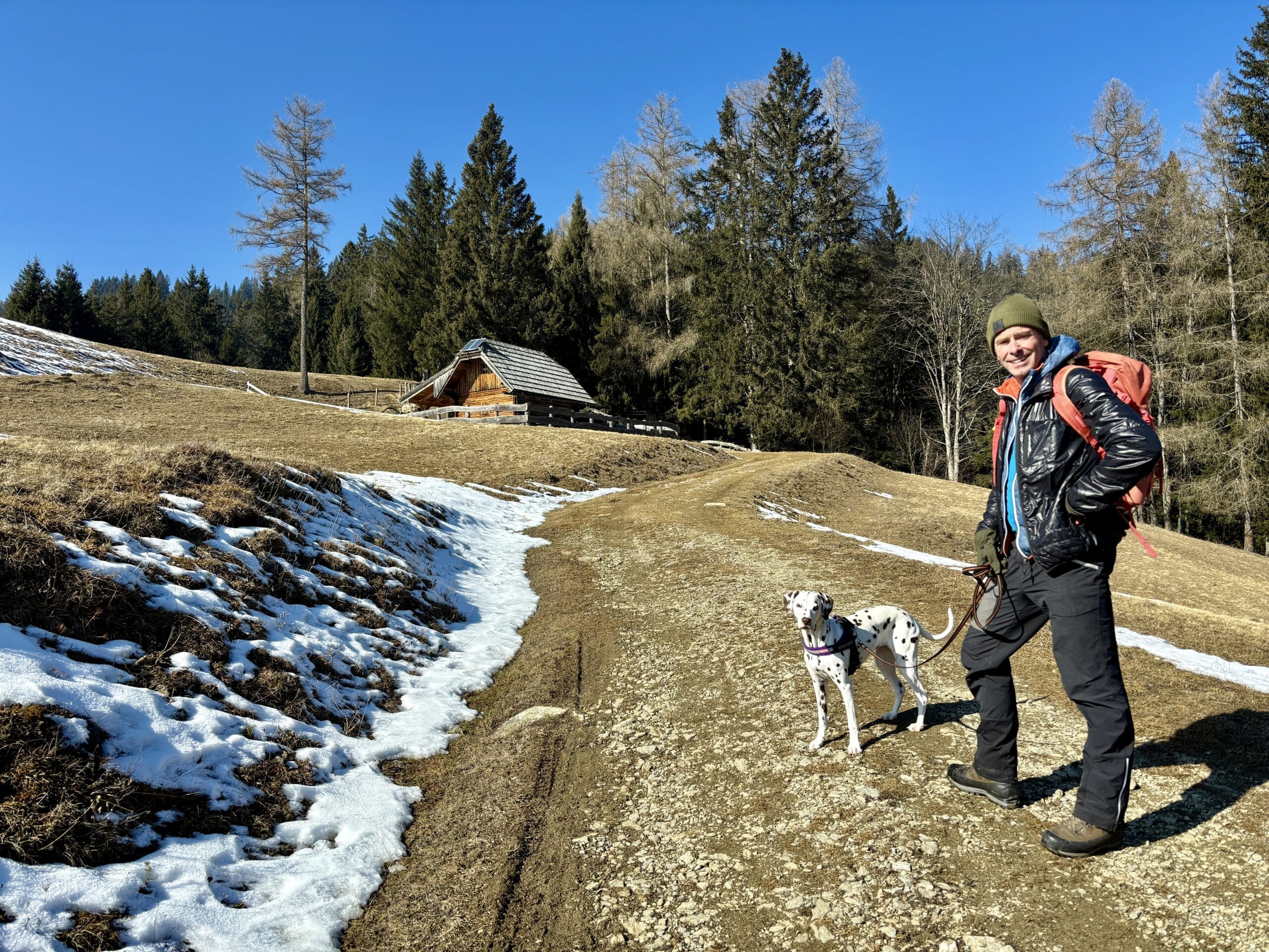 Mann wandert mit Hund auf Alm, im Hintergrund sieht man Almhütte.
