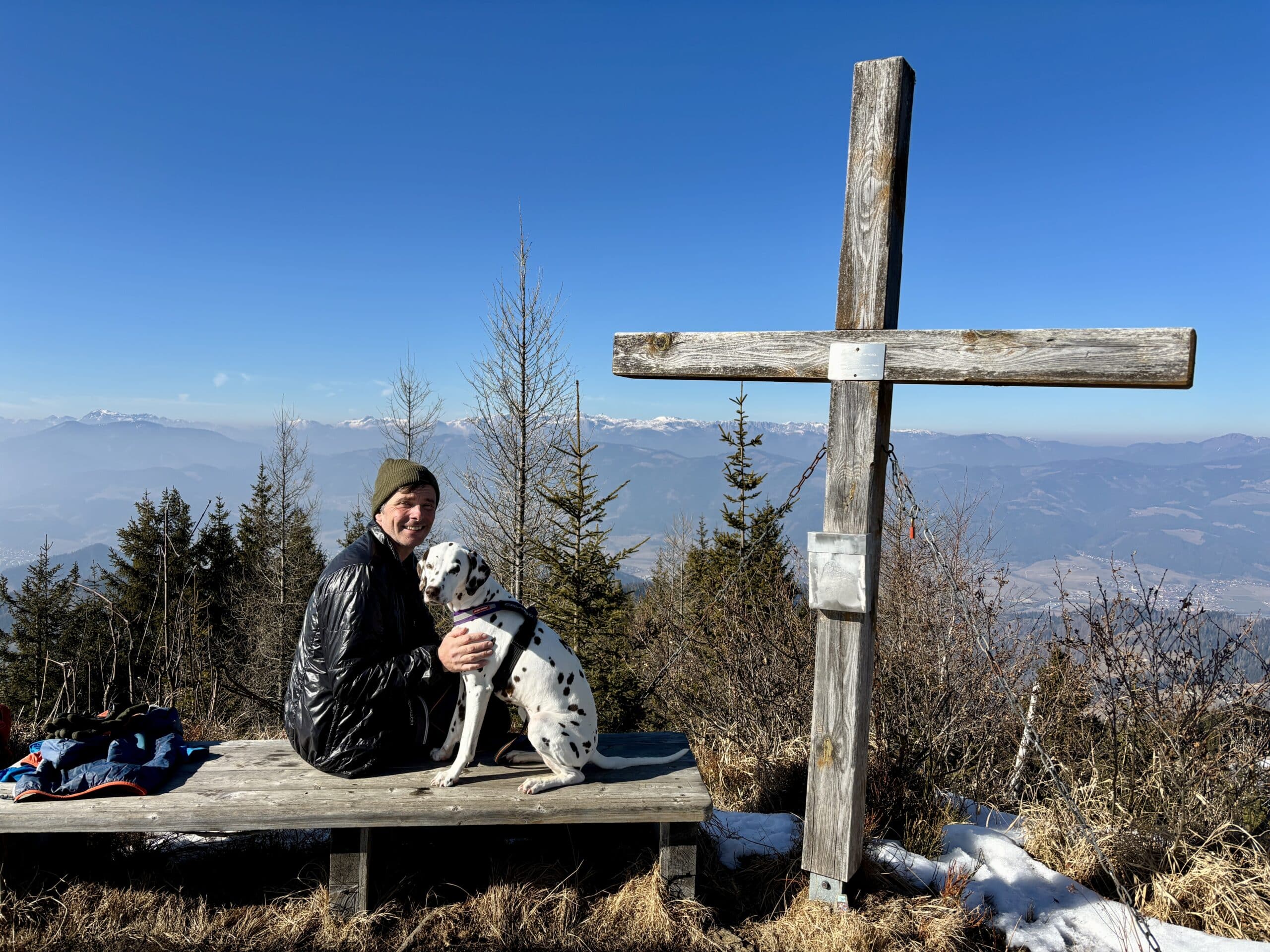 Mann sitzt mit Hund (Dalmatiner) auf Bank neben Gipfelkreuz