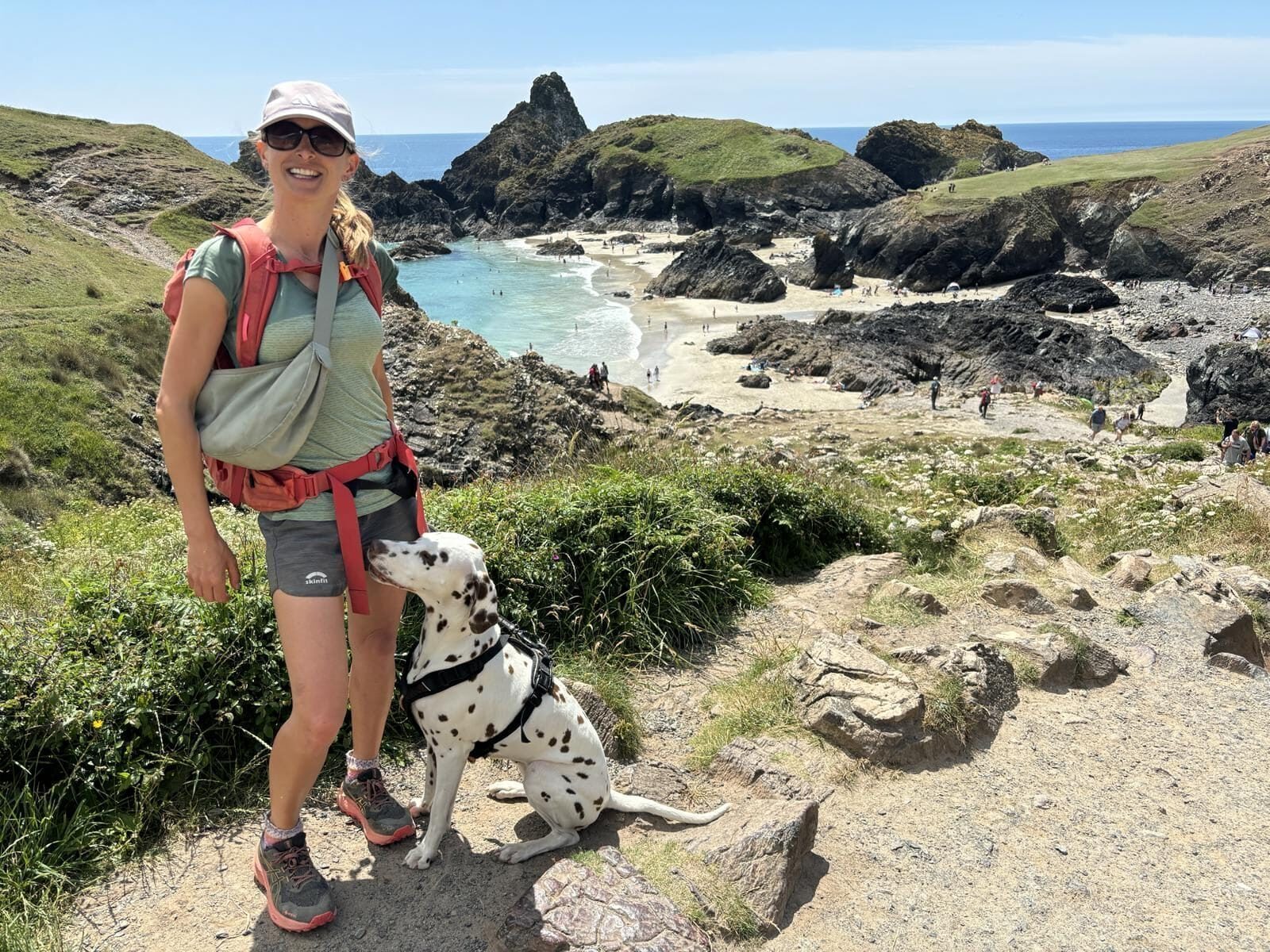 Frau steht mit Hund (Dalmatiner) in Wanderkleidung über Bucht; weißer Strand, schwarze Felsen