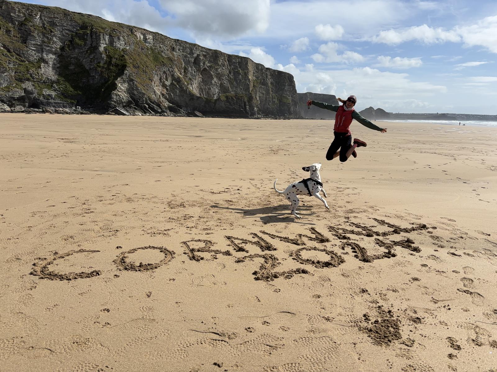 Frau springt an einem breiten, von Felsen umrahmten Strand; im Sand steht Cornwall 2024; Hund schaut zu