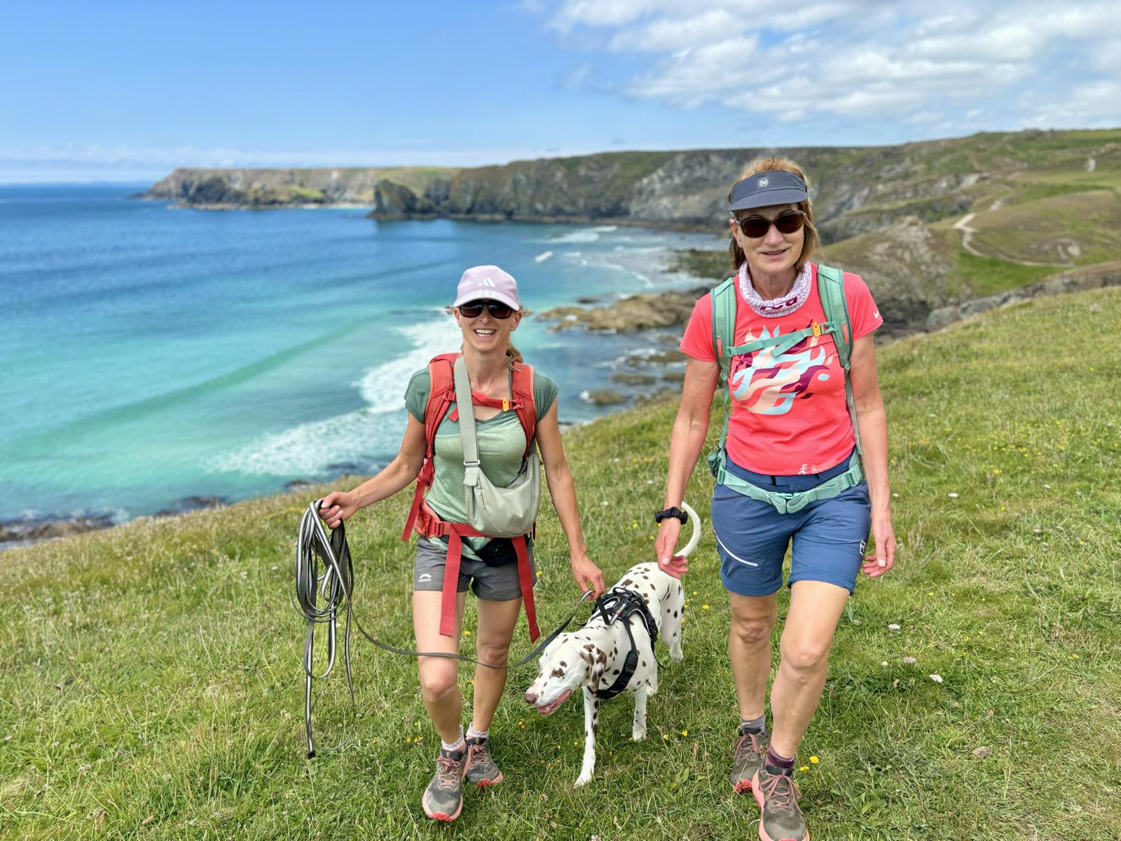 Zwei Frauen wandern durch Wiese mit Hund an einem Küstenwanderweg