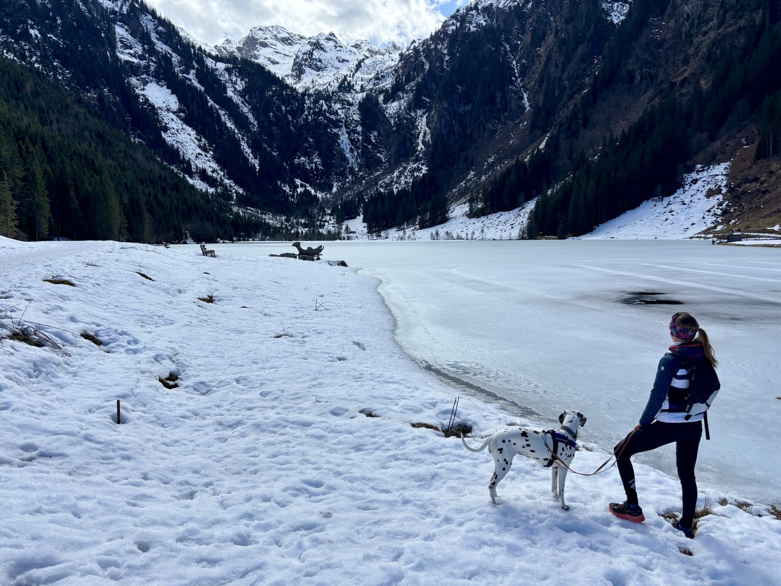 Frau steht mit Hund an schneebedecktem Seeufer in den Bergen.