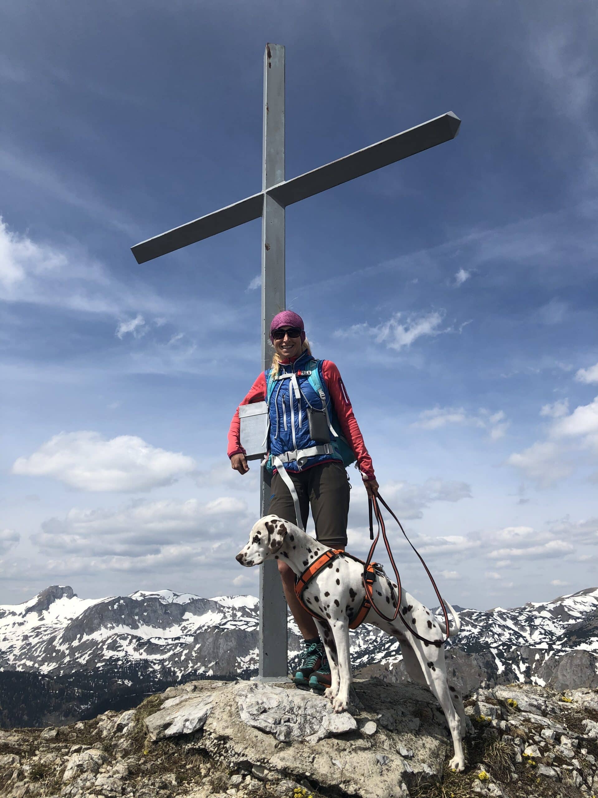 Frau steht mit Hund bei Gipfelkreuz aus Eisen