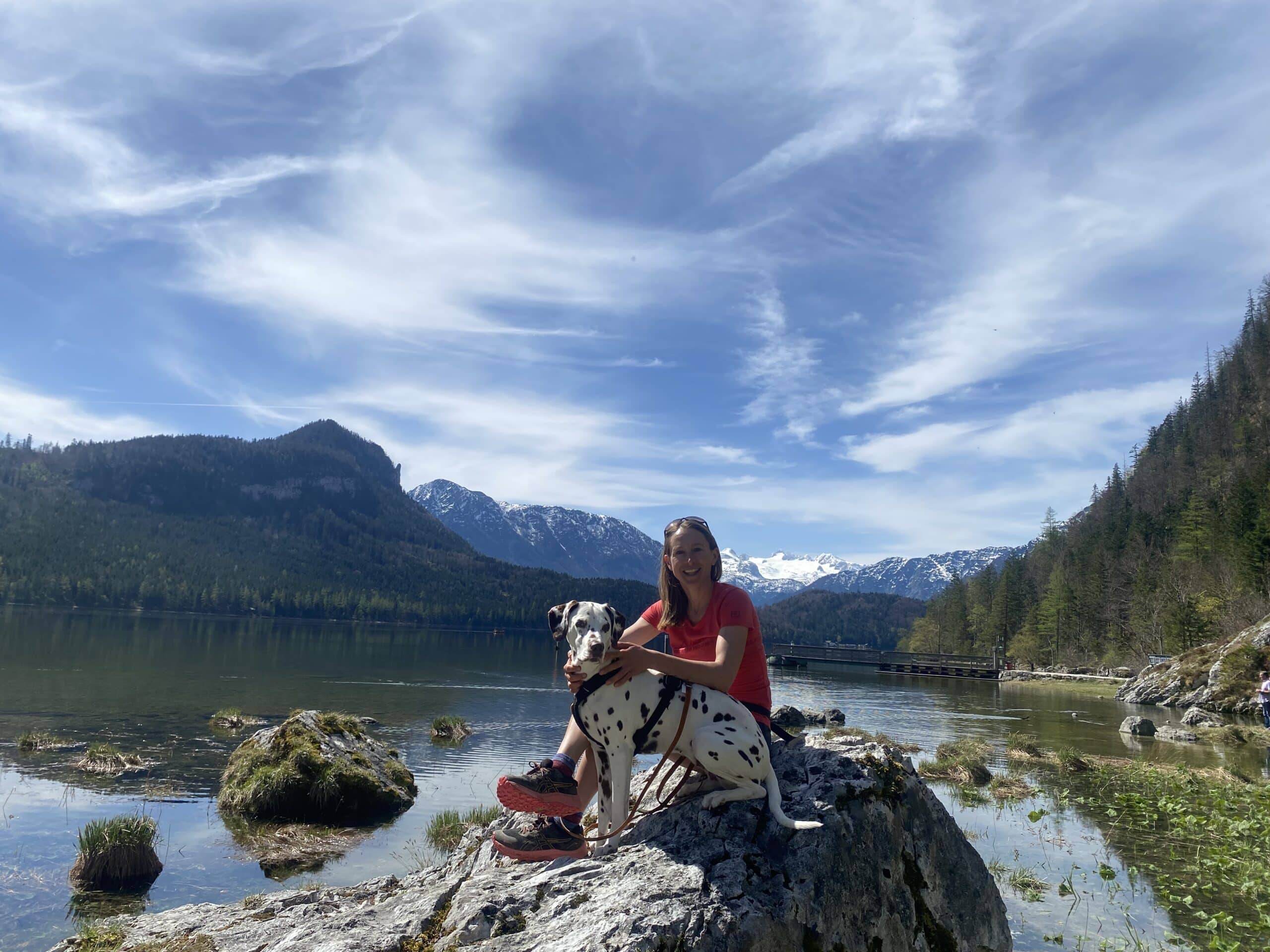 Frau sitzt mit Hund auf einem Felsen an einem See