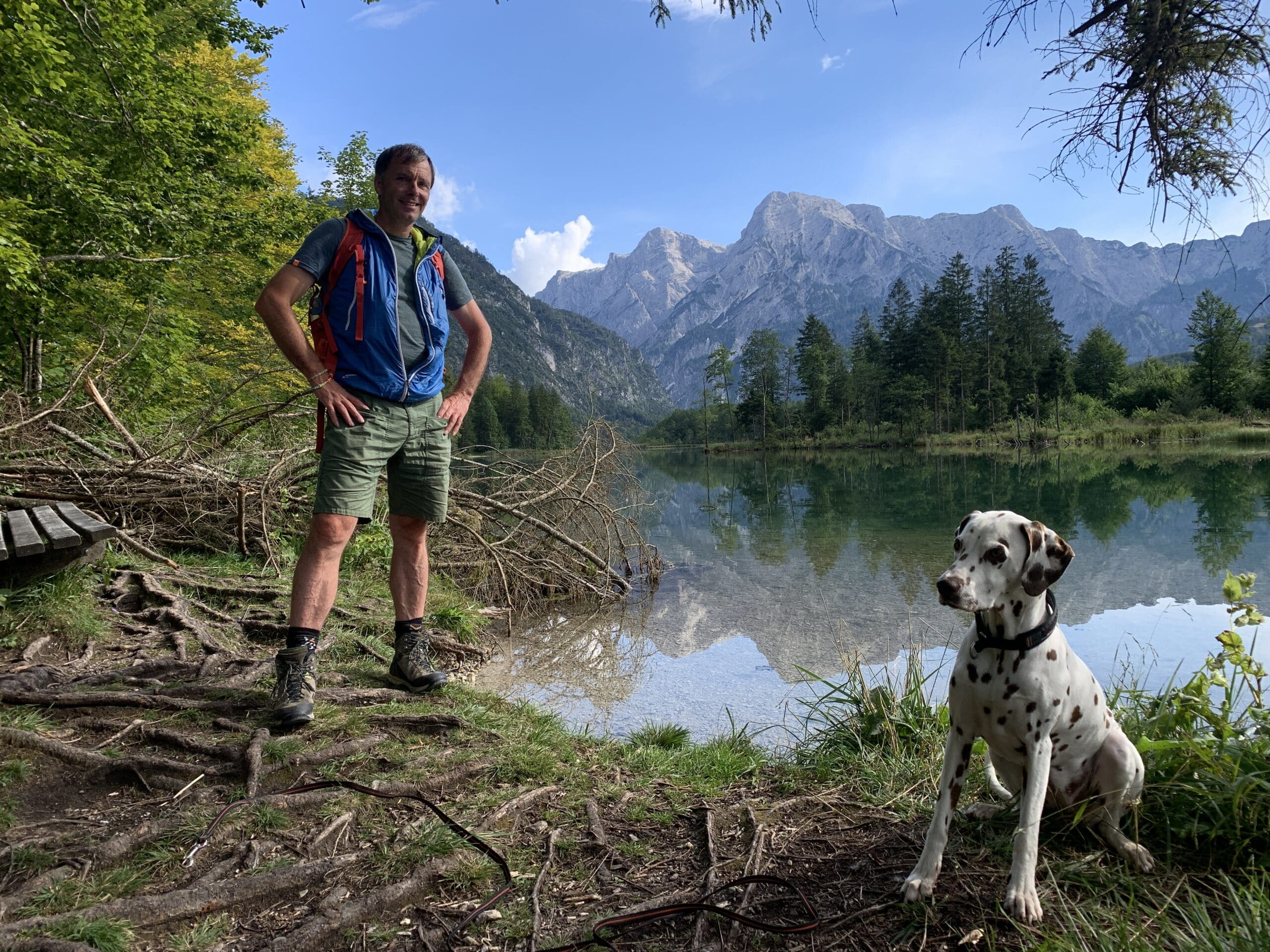 Mann steht mit Hund am Ufer eines Sees, umrahmt von schroffen Bergen