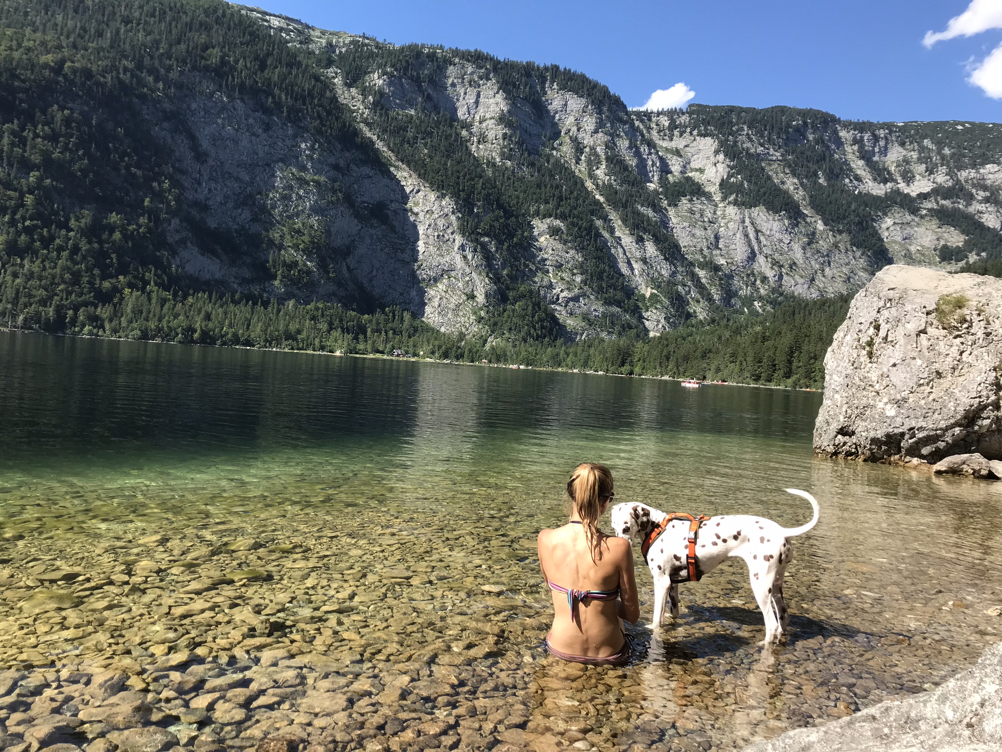 Frau sitzt im Wasser in einem glasklaren Bergsee. Hund steht daneben im Wasser