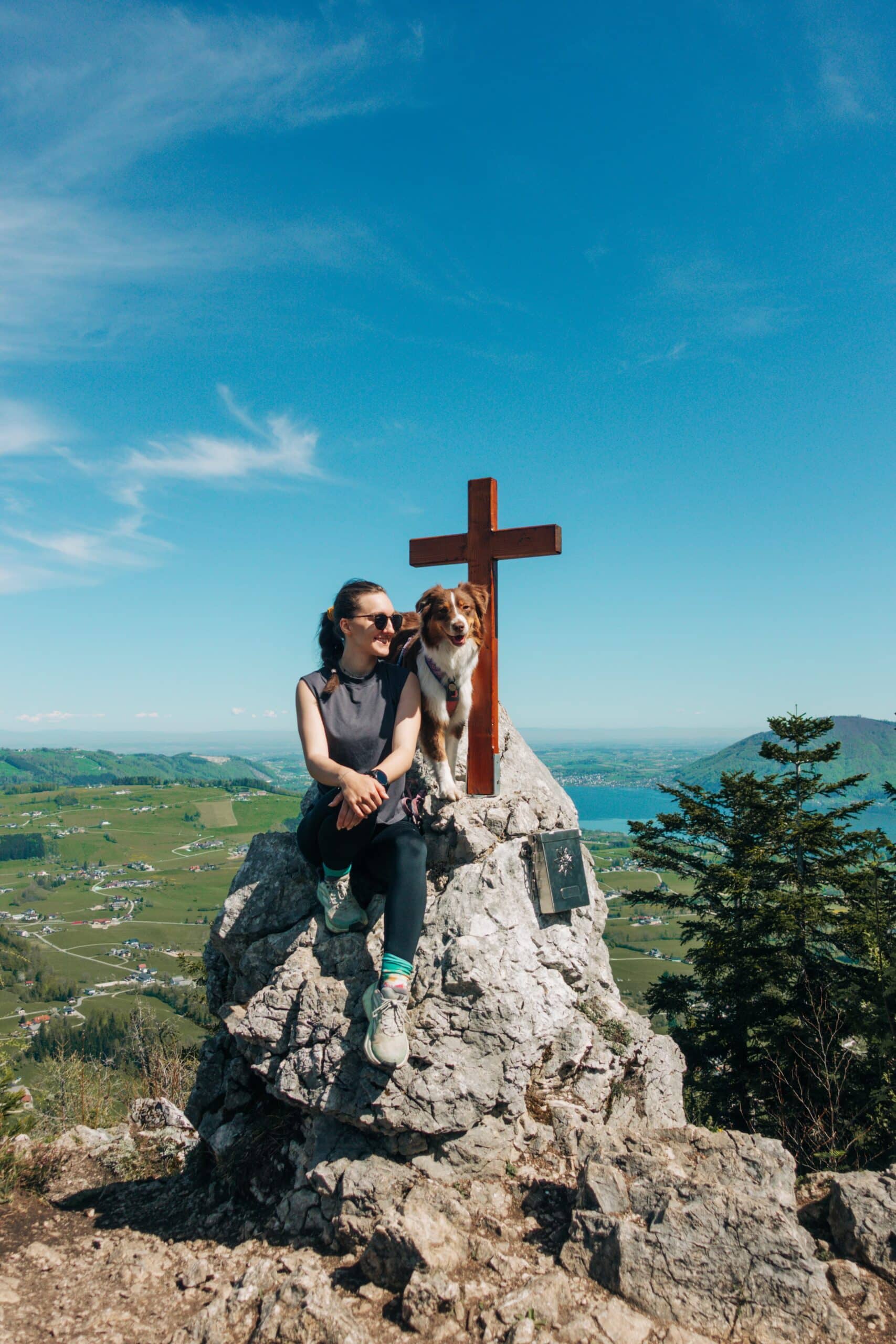 Frau sitzt mit Hund bei Gipfelkreuz auf Felsen